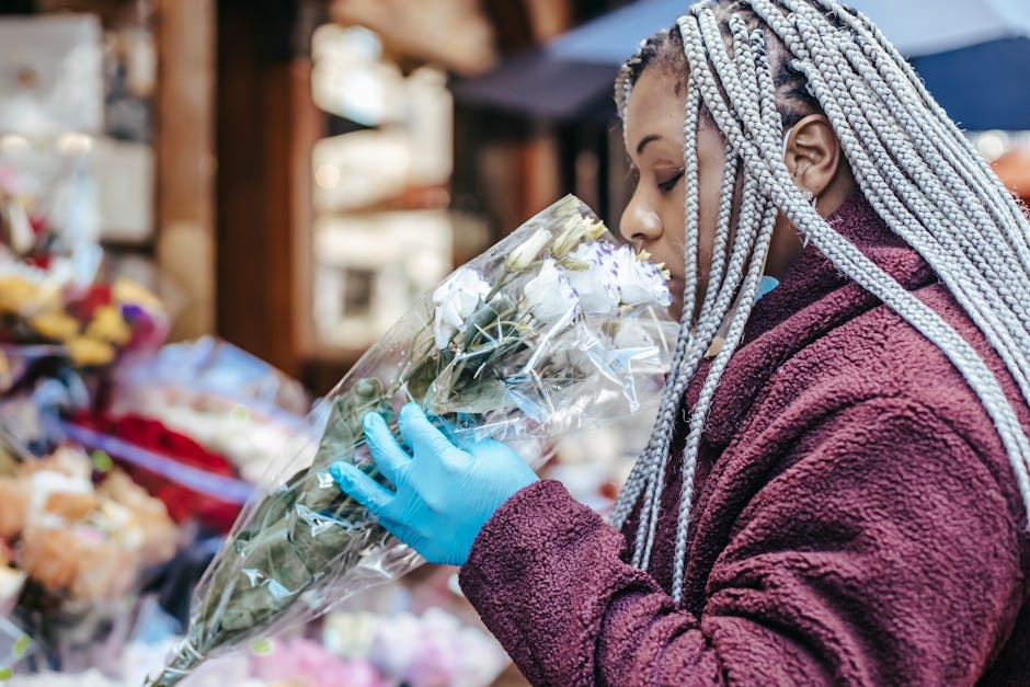 A woman with long, braided, silver-gray hair, wearing a burgundy fleece jacket and bright blue gloves, is inspecting a bouquet of white roses wrapped in clear cellophane at an outdoor flower stall. Th