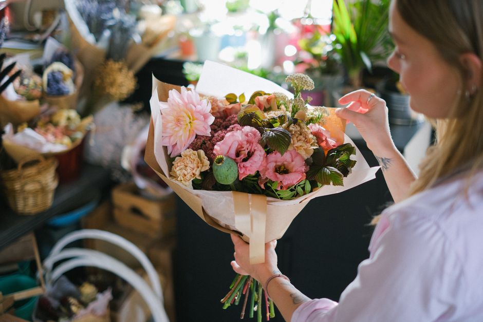 A woman with long, light brown hair is seen from the side, holding a large, vibrant bouquet of fresh flowers wrapped in white and beige paper with a satin ribbon. The bouquet features soft pink and pe