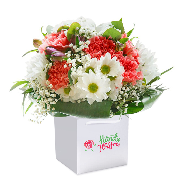 Nestled in a stylish, sturdy white square gift bag, this bold and uplifting Creekmouth bouquet promises freshness and joy from the moment it arrives. A generous arrangement of soft coral and rich peach carnations forms the plush heart of the display, their lavish layers of ruffled petals softening the eye and inviting a tactile appreciation. Among them, elegant white daisies tipped with radiant yellow-green centres stand out, ushering in a wave of cheerful brightness. Light, floating sprays of baby's breath interlace through the flowers, introducing an ethereal texture that brings effortless delicacy. On the left, subtle traces of new purple buds signal a continuing story of growth, while a well-crafted surround of deep green, glossy leaves provides both structure and sumptuous colour-echoing the leafy perimeters of Creekmouth's riverside greenery. The practical white gift bag, with reinforced ribbon handles, allows for convenient gifting or local delivery, while the whole ensemble is displayed on a clean white background to highlight every; detail. This full, rounded bouquet is as lively as Creekmouth itself, suitable for celebrating milestones or simply spreading a bright gesture throughout the area.